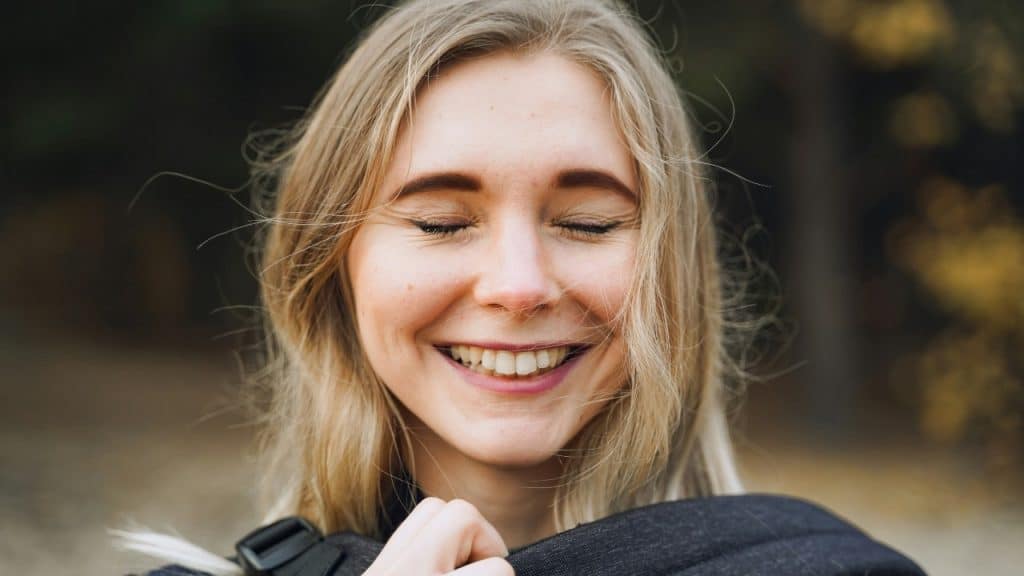 A blonde woman smiles with her eyes closed while holding the straps of a black backpack.