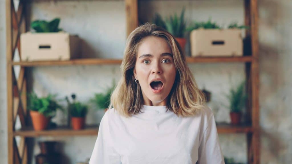 A woman in a white shirt looks forward with a wide-eyed, open-mouthed expression of surprise.