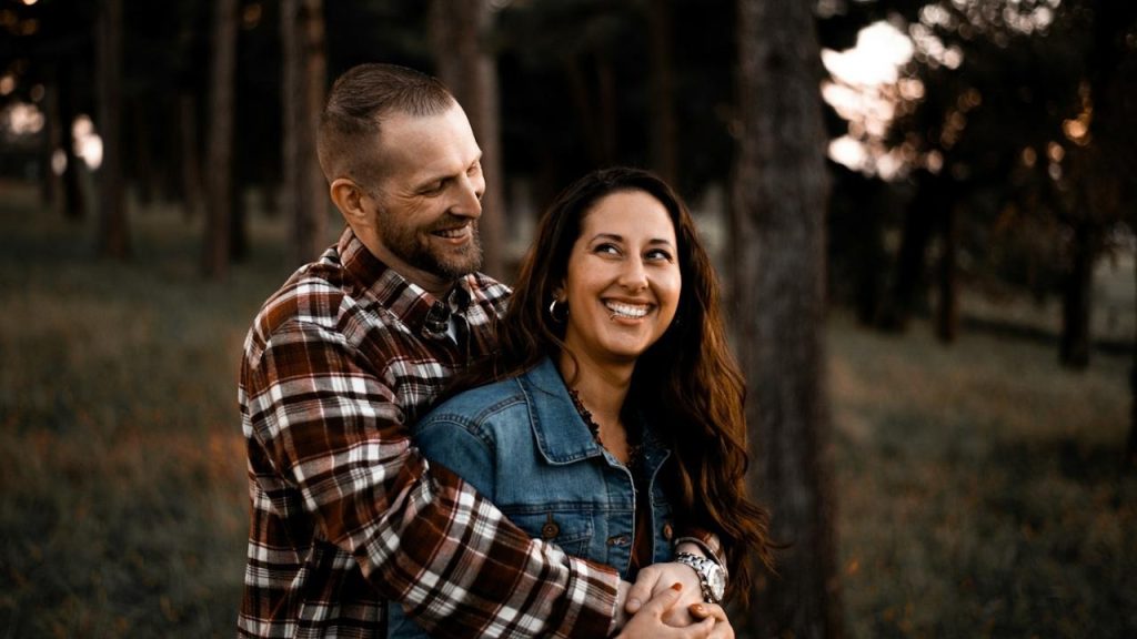 A smiling man embraces a woman from behind in a forest setting.