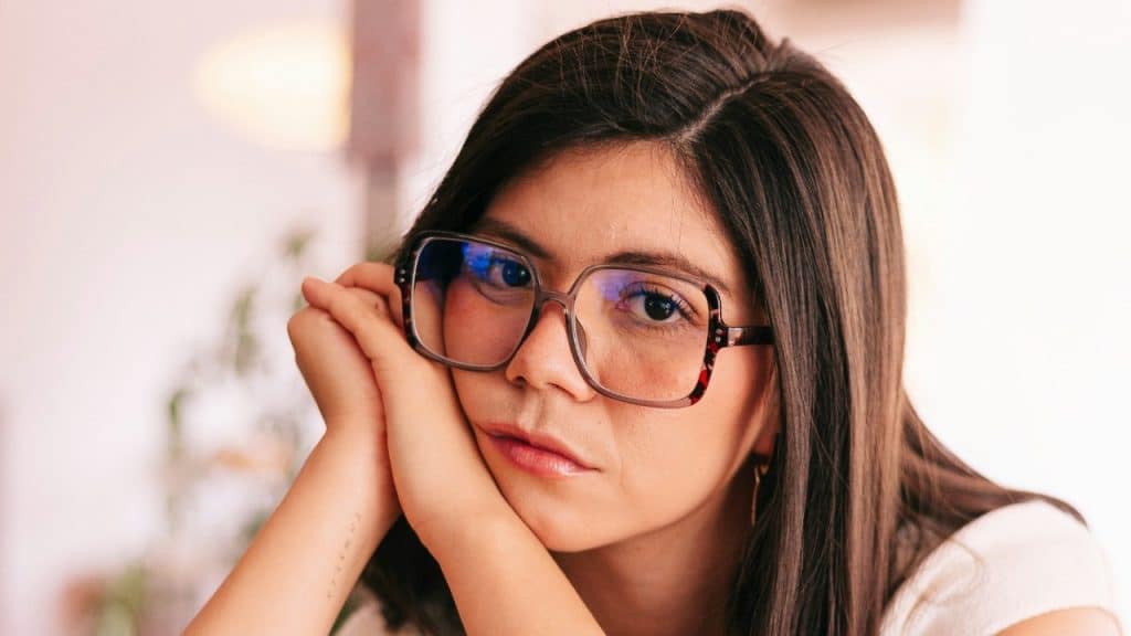 A woman wearing large tortoiseshell glasses rests her chin on her hands and looks at the camera.