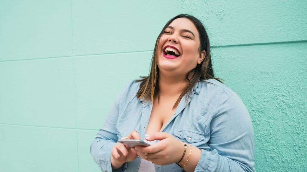 A woman in a blue denim shirt laughs joyfully while holding a smartphone against a teal wall.