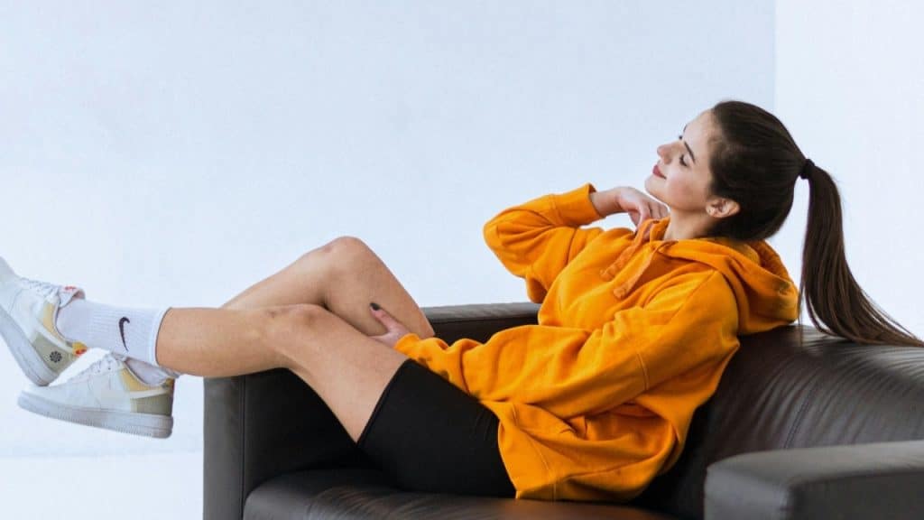 A woman in an orange hoodie and black shorts reclines on a dark leather sofa.