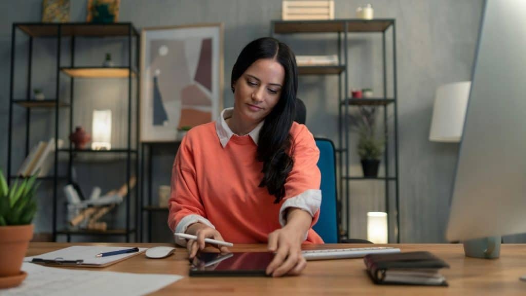 A woman in a coral sweater sits at a desk while using a tablet and stylus.