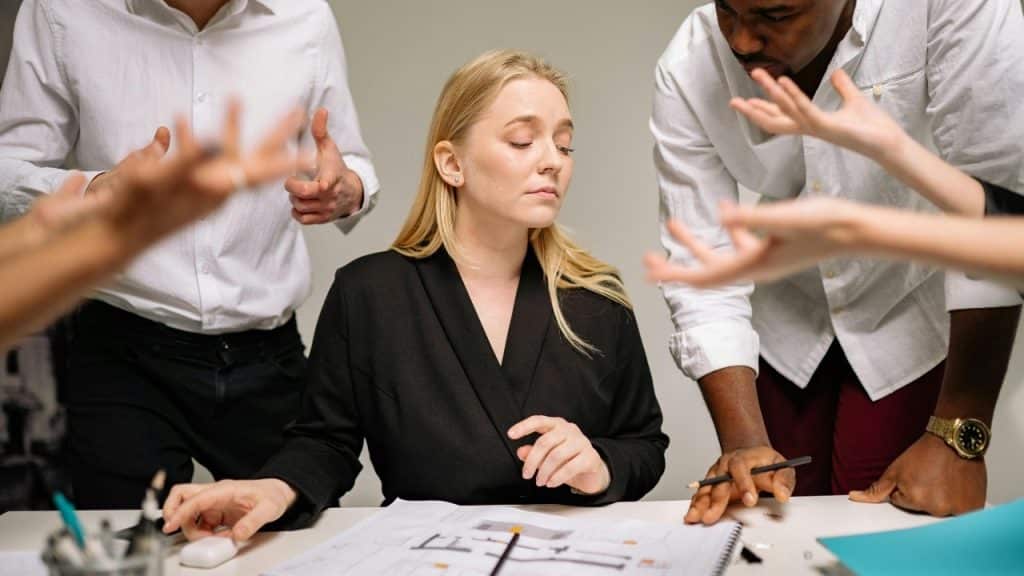A blonde woman sits calmly at a desk while several people gesture around her with frustration.