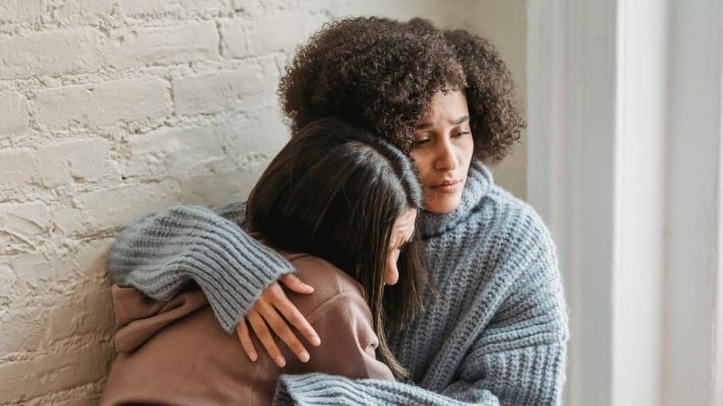A woman with curly hair wearing a grey sweater embraces another person against a brick wall.