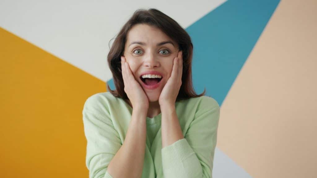 A woman in a light green sweater holds her cheeks with a surprised, open-mouthed expression.