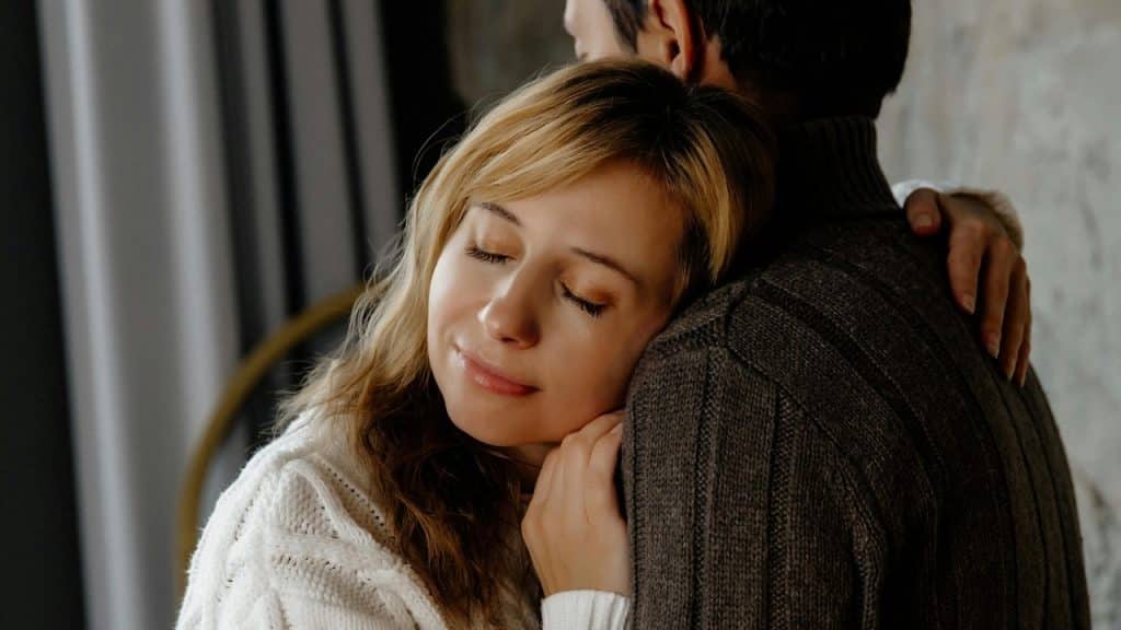 A blonde woman with eyes closed leans her head against a person in a brown sweater.