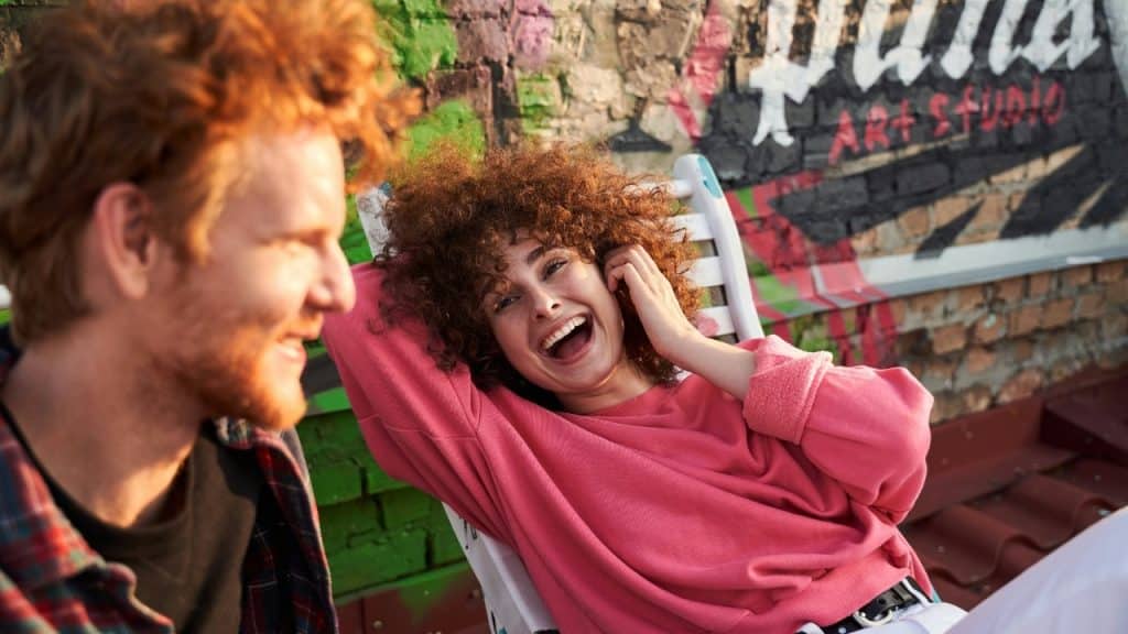 A woman with curly hair laughs while reclining in a chair outdoors next to a man.