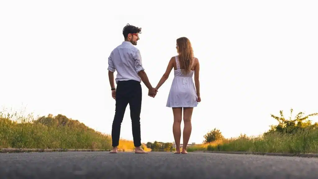 A man and a woman stand barefoot on a paved road, holding hands and looking toward a bright, sunlit horizon bordered by tall grass.