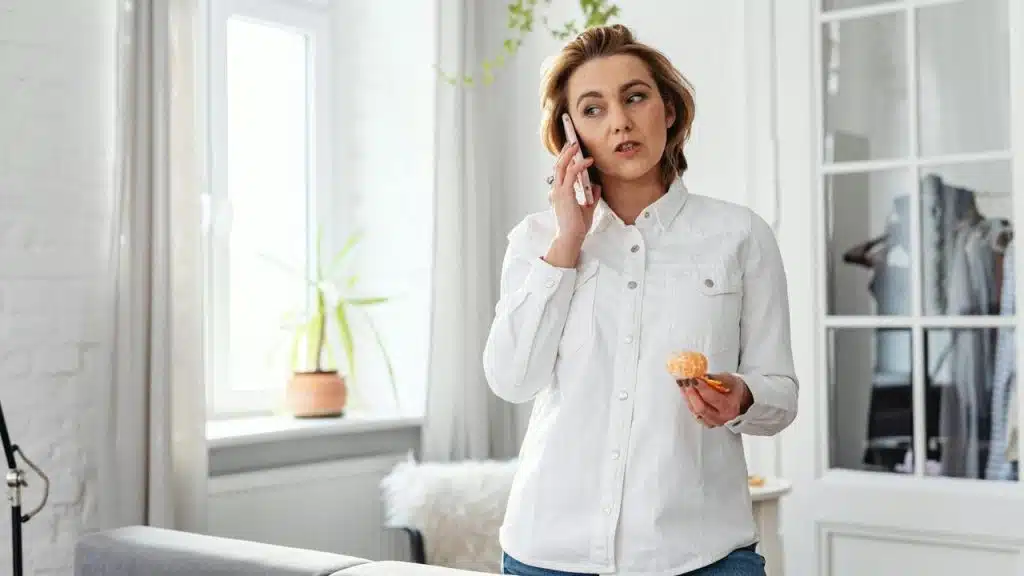 A woman talks on her phone while holding a peeled orange at home.