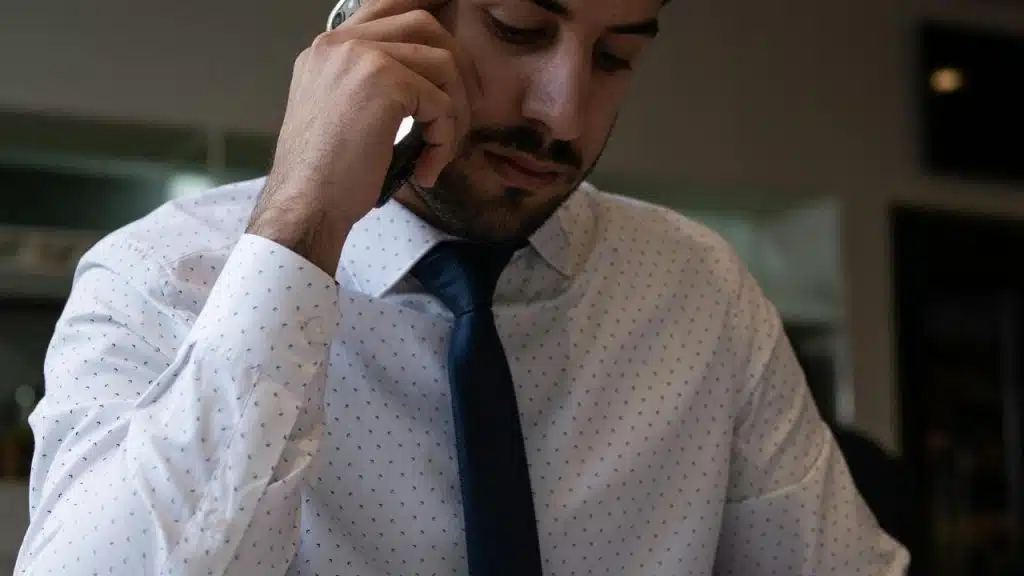 A man in a dress shirt and tie talking on a phone while looking down.