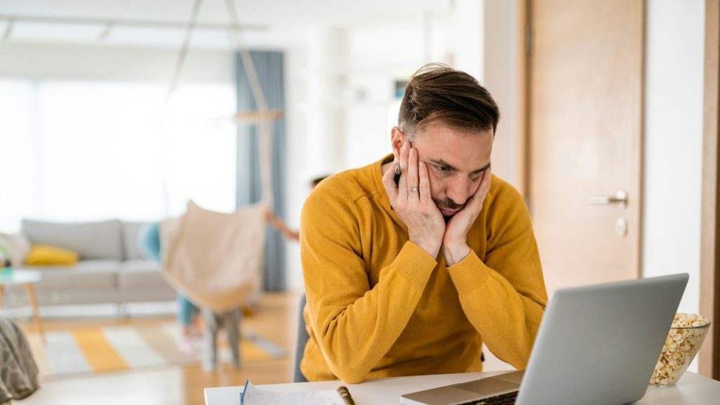 A tired man holding his face in his hands is working on the laptop.