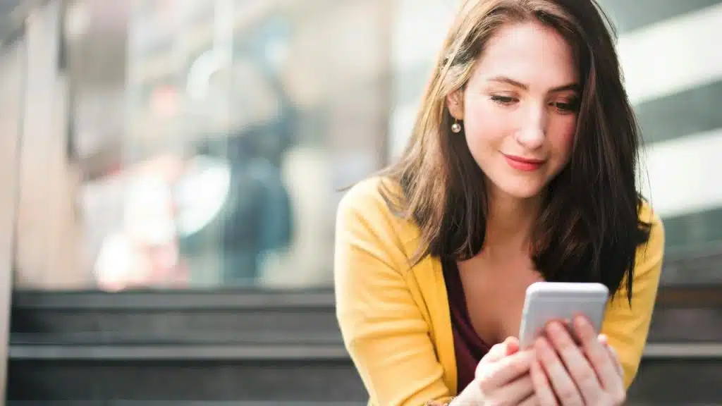 A woman in a yellow cardigan looking at her smartphone indoors.