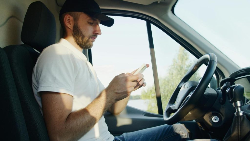 A man sitting in the driver’s seat of a vehicle while using a smartphone.