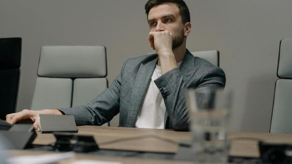 A man in a blazer sitting at a table with his hand over his mouth.