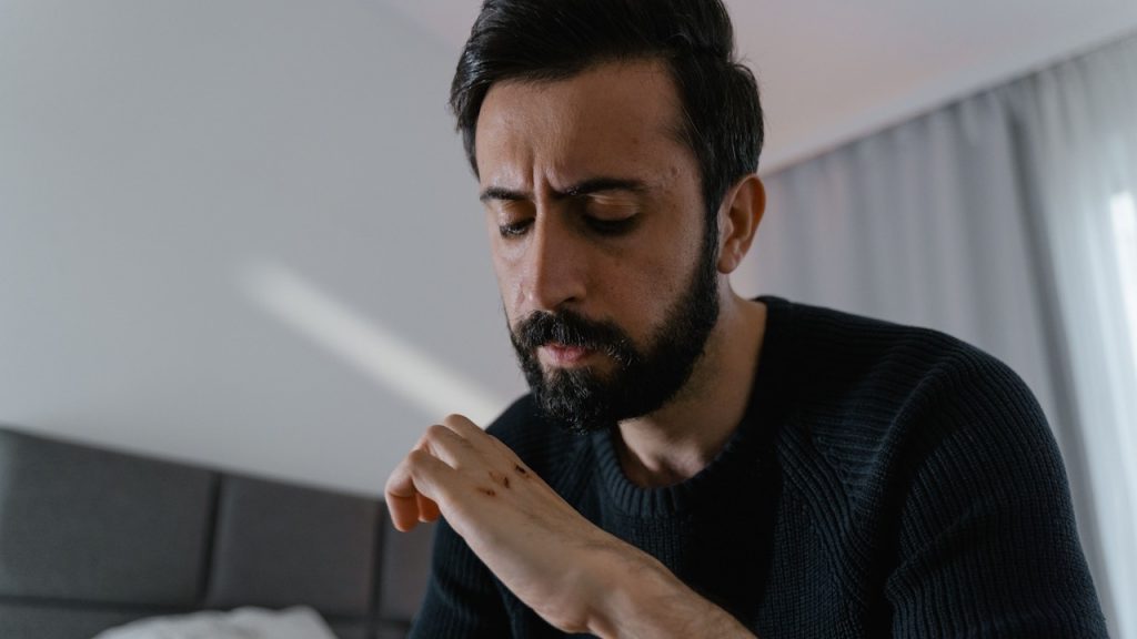 A bearded man sitting on a bed and looking at his hand.