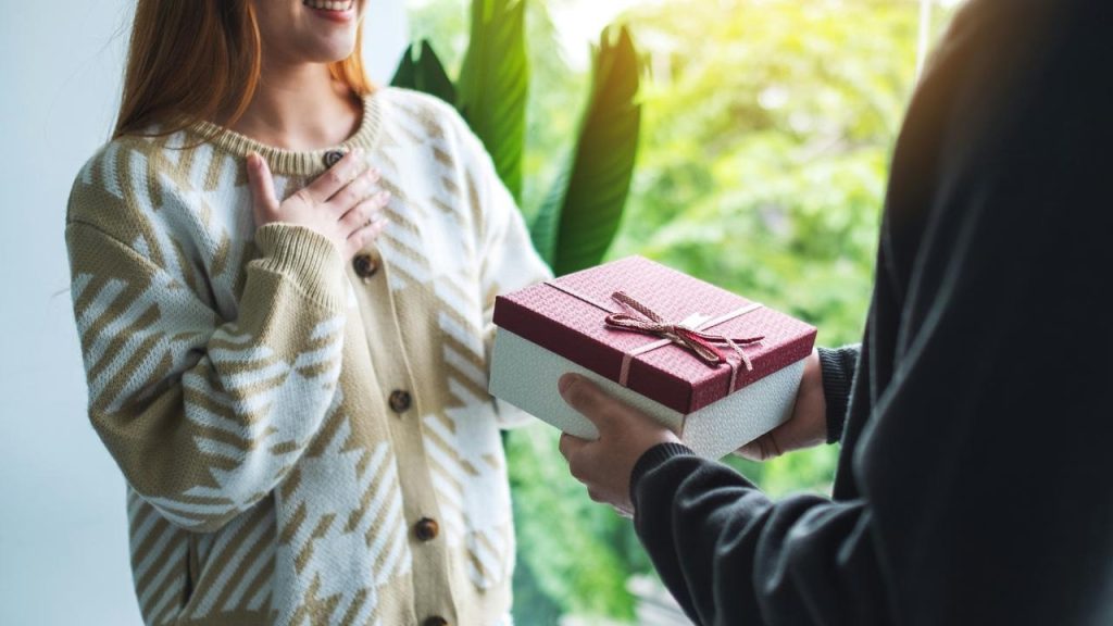 A person handing a wrapped gift box to a smiling woman indoors near a window.