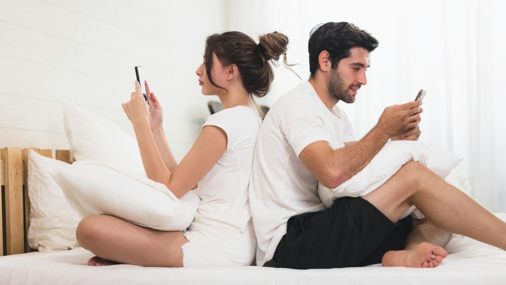 A couple sitting with their backs to each other on a bed and using their phones.