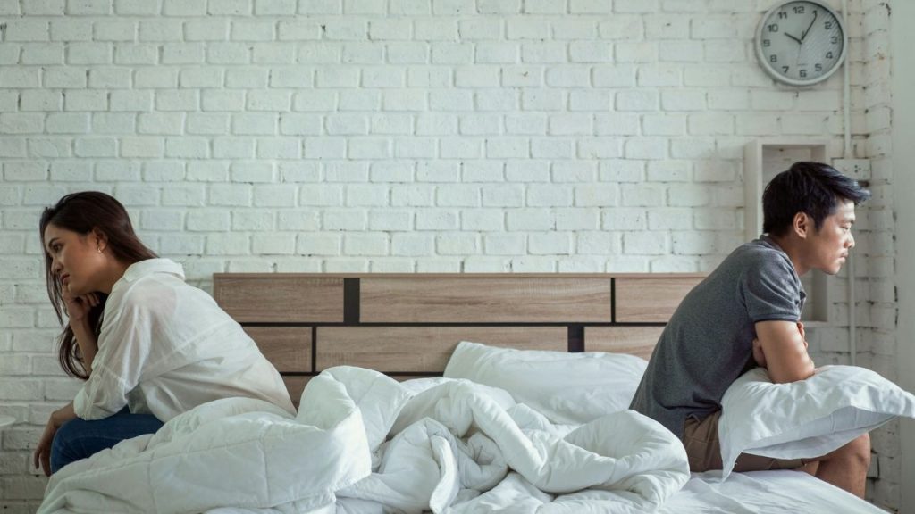A man and woman sit on a bed with their backs facing toward each other.