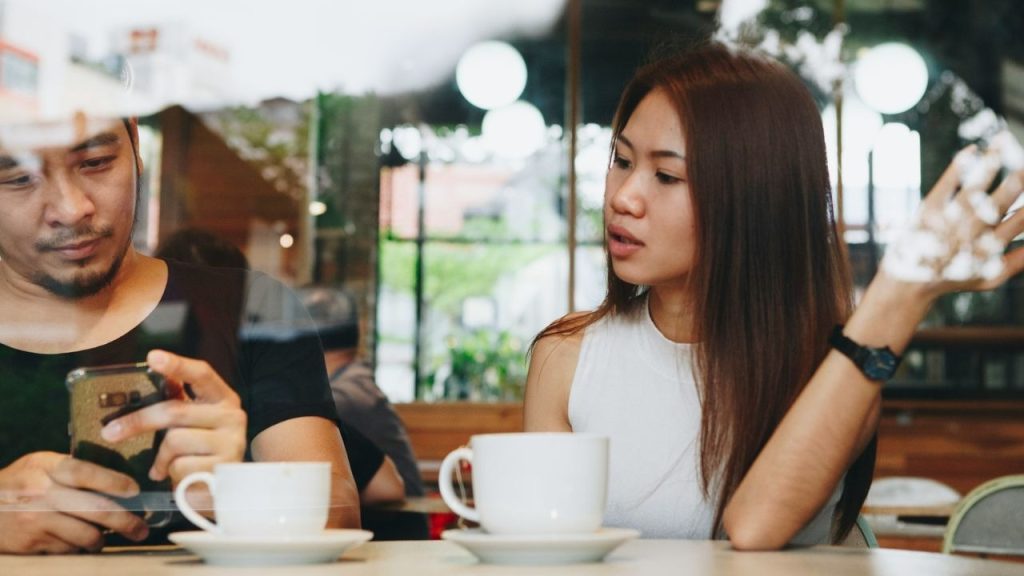 A woman speaks to a man who is looking down at his smartphone in a cafe.