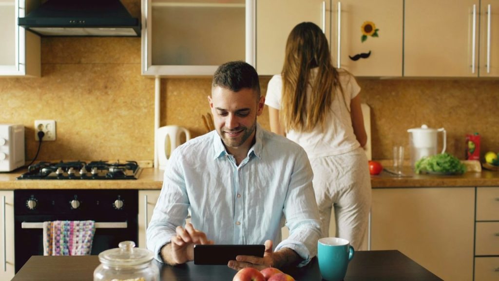 A man uses a tablet at a table while a woman works in the kitchen background.