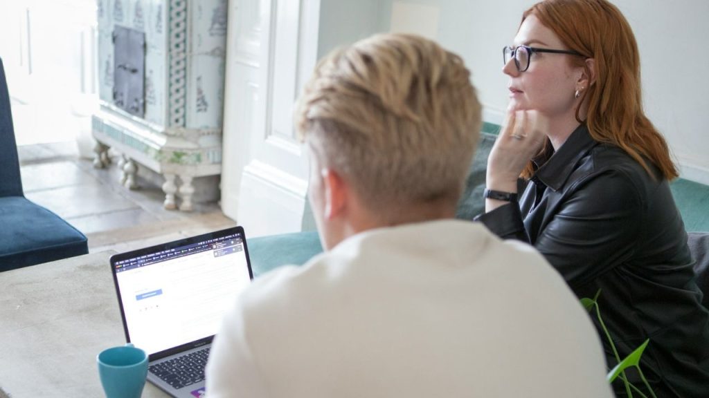 A person uses a laptop while another person sits nearby looking away into the distance.