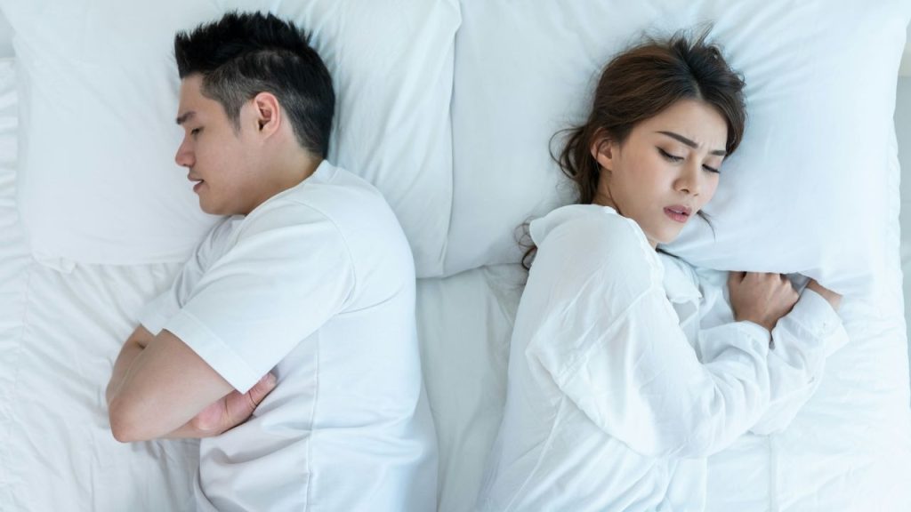 A man and woman lie in bed facing away from each other with eyes closed.