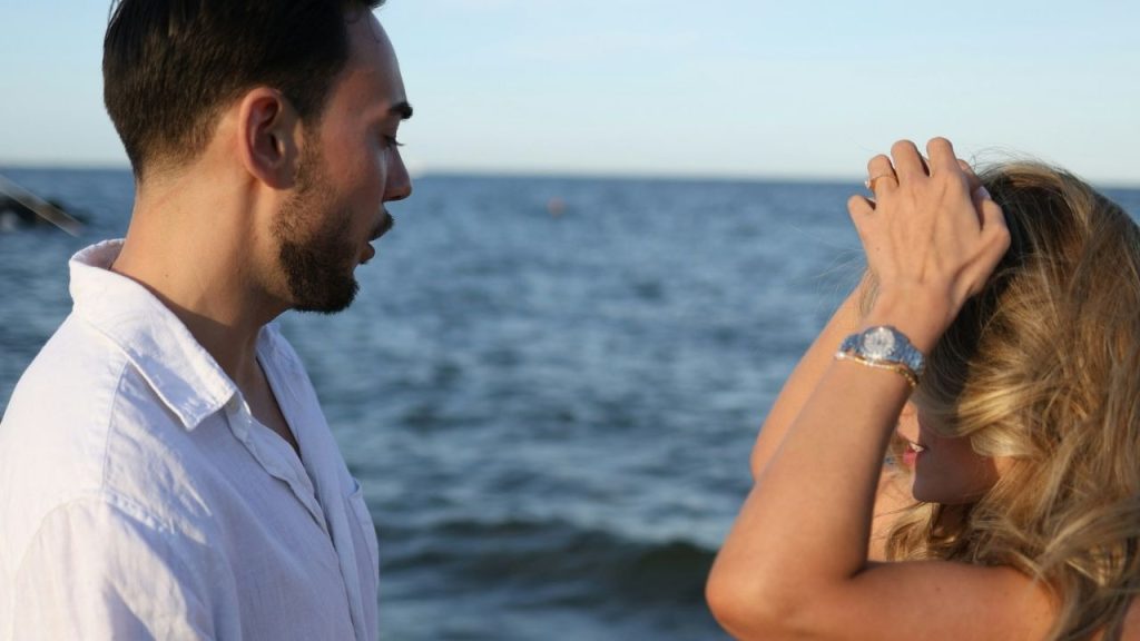 A man and woman stand near the ocean while she adjusts her blonde hair.