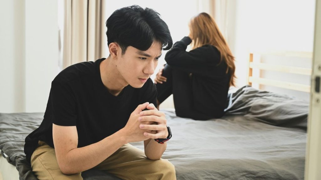 A man sits on a bed with hands clasped, while a woman sits facing away behind him.