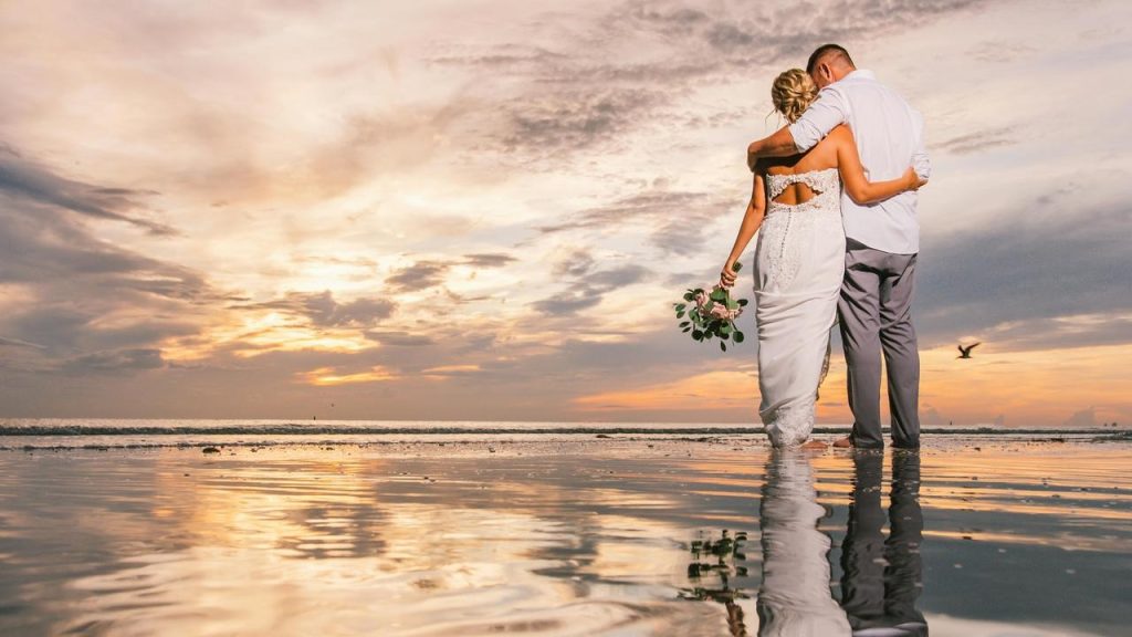 A couple embracing on a beach at sunset, reflected in wet sand.