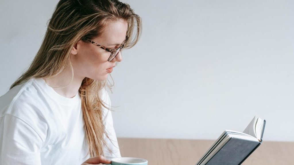 A woman wearing glasses reading a book while holding a cup.