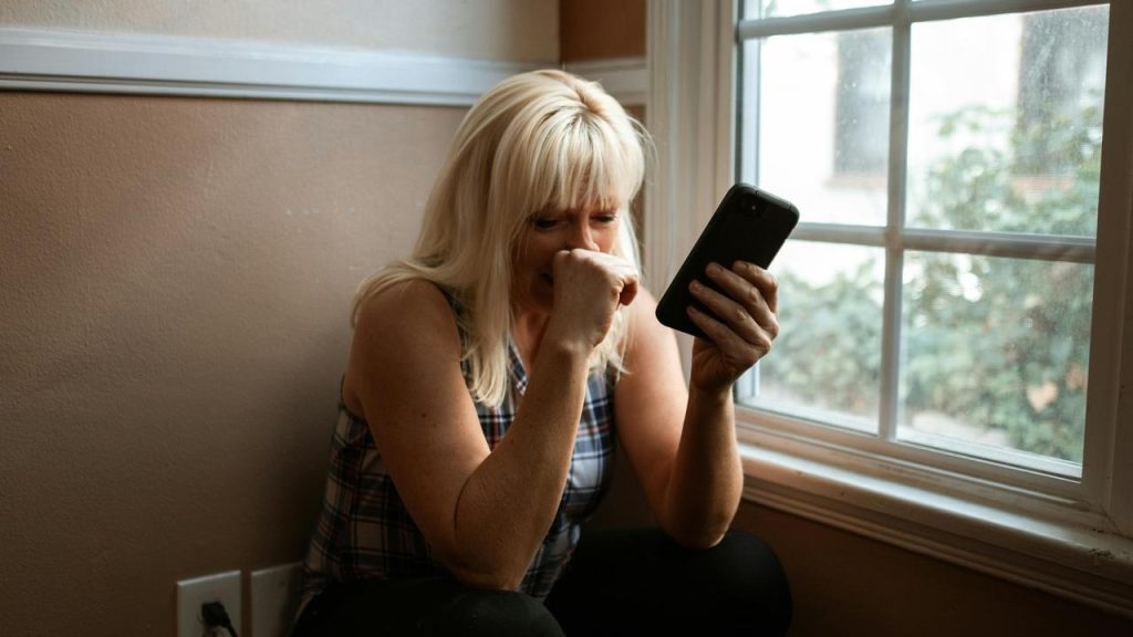 A woman by a window looking at her phone with a distressed expression.
