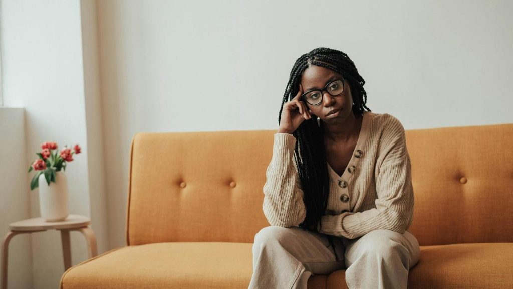 A woman wearing glasses sitting on a couch with her hand resting against her temple.