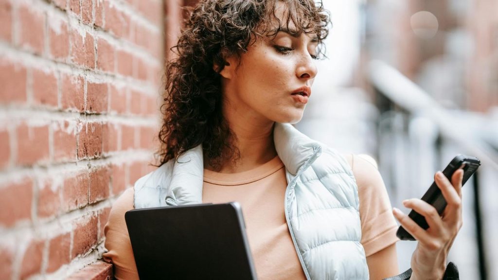 A woman standing by a brick wall holding a laptop and looking at her phone.