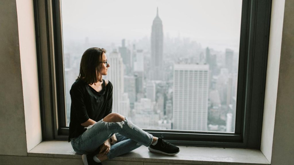 A woman sits by a large window, gazing out at a city skyline with tall buildings in the distance.