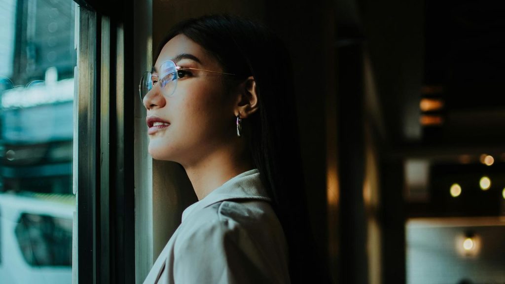A woman wearing glasses standing by a window and looking outside.