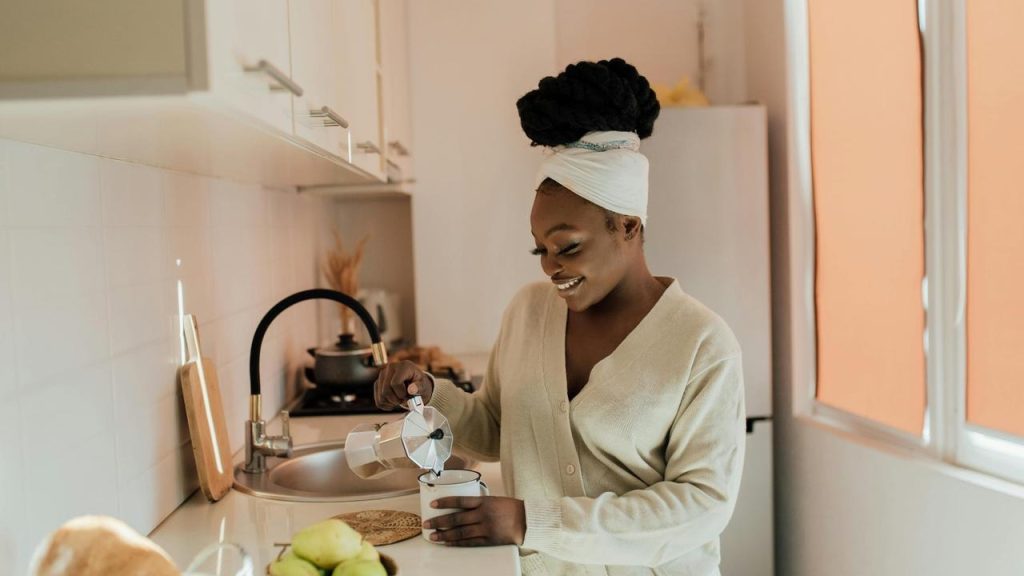 A woman stands in a bright kitchen, smiling as she pours coffee into a cup.