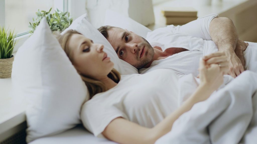 A couple lying in bed while the man reaches for the woman, who looks a bit sad.