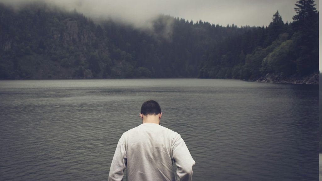 A man stands before a lake surrounded by trees.