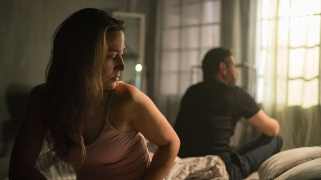 A woman and man sitting with their backs to each other and not talking while they sit on a bed.