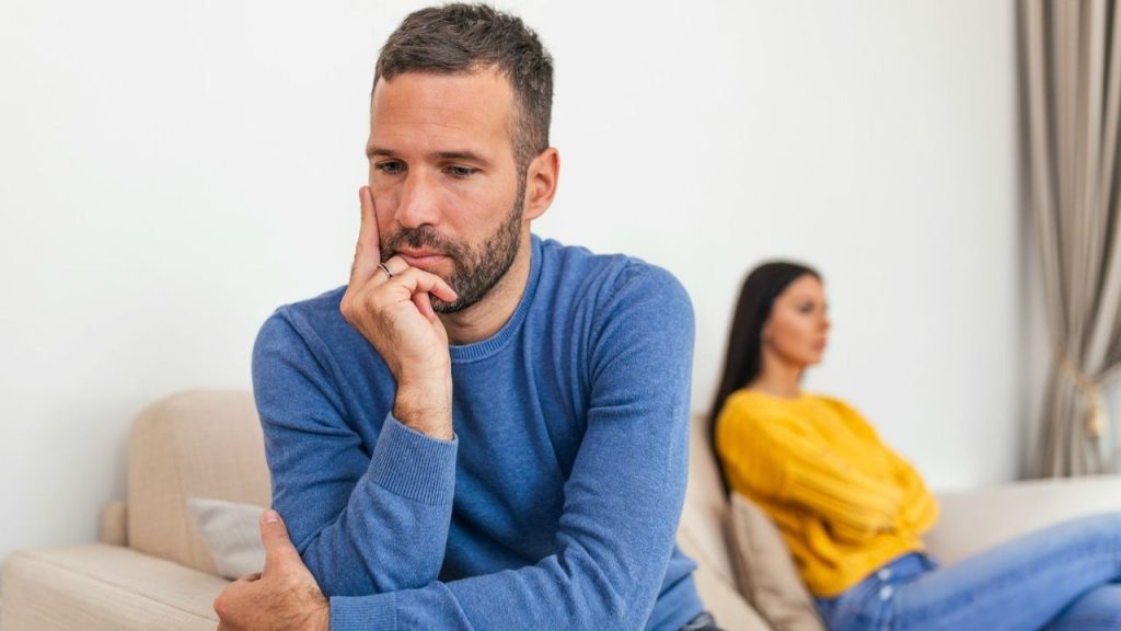 A man in a blue sweater sits with a pensive expression, while a woman in yellow is blurred in the background.