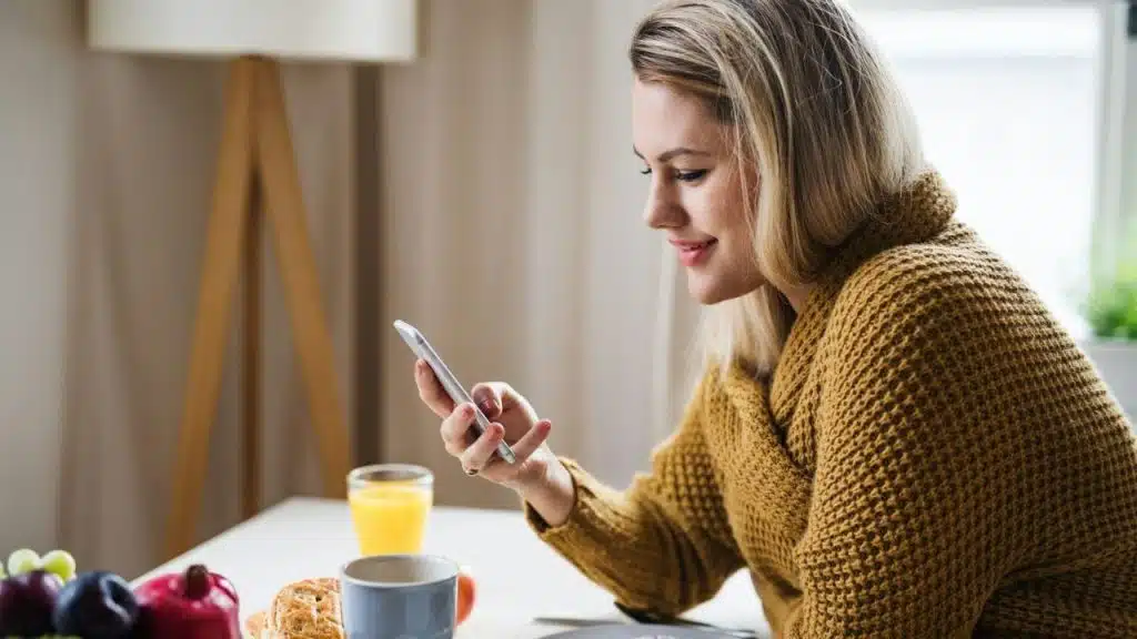 A smiling woman in a yellow sweater is looking at her phone at a breakfast table.