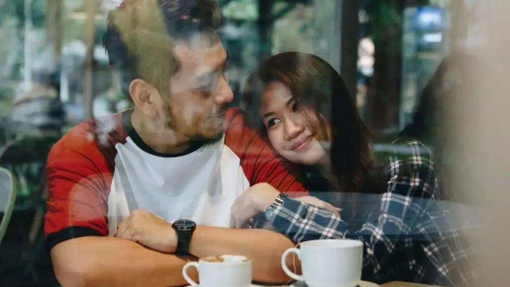 A woman rests her head on a man's shoulder at a cafe table, looking at him with a smile.