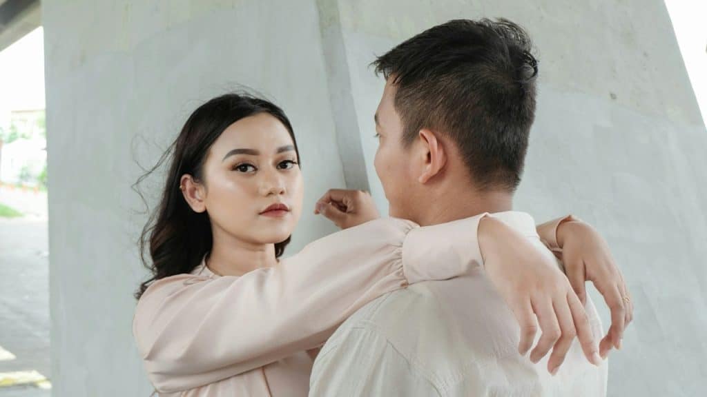 A woman with an inscrutable expression as she looks at the camera while resting her arms on a man’s shoulders.