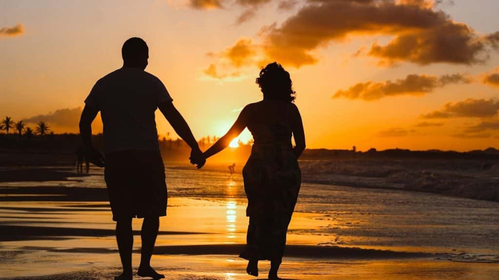 A silhouette of a couple walking while holding hands down a beach.
