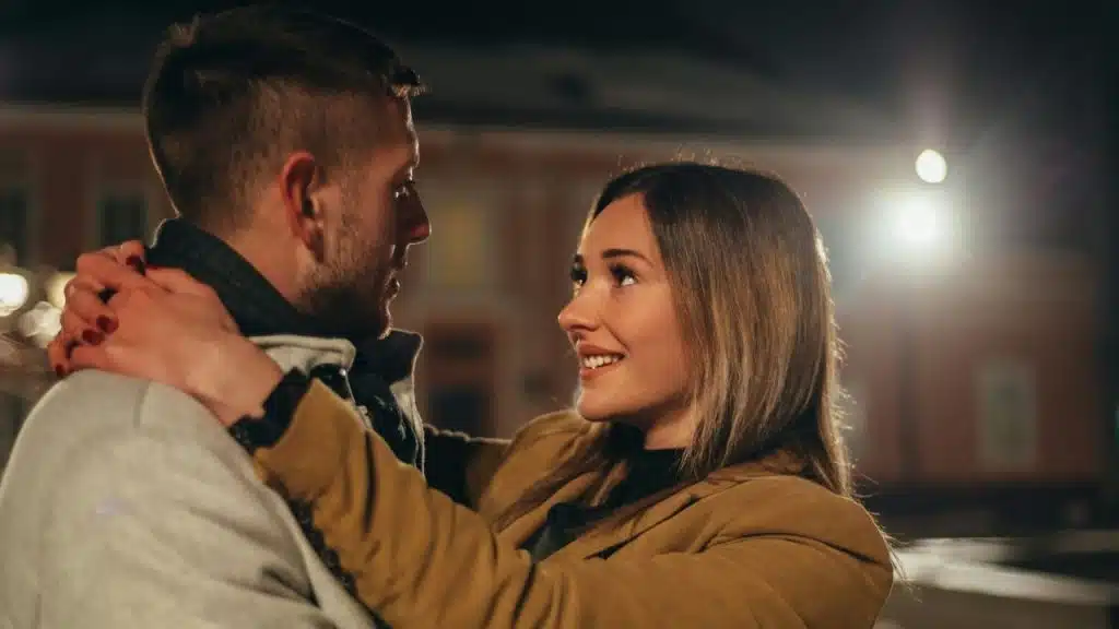 A man and a woman embrace outdoors at night, looking at each other with smiles.