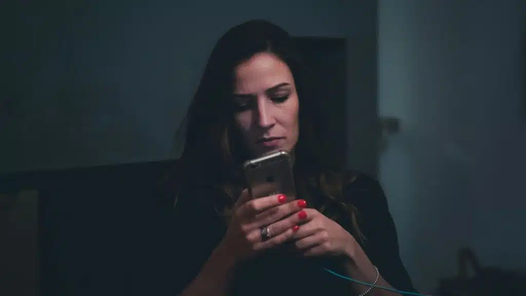 A woman with red nail polish looks intently at her phone in a dimly lit room.