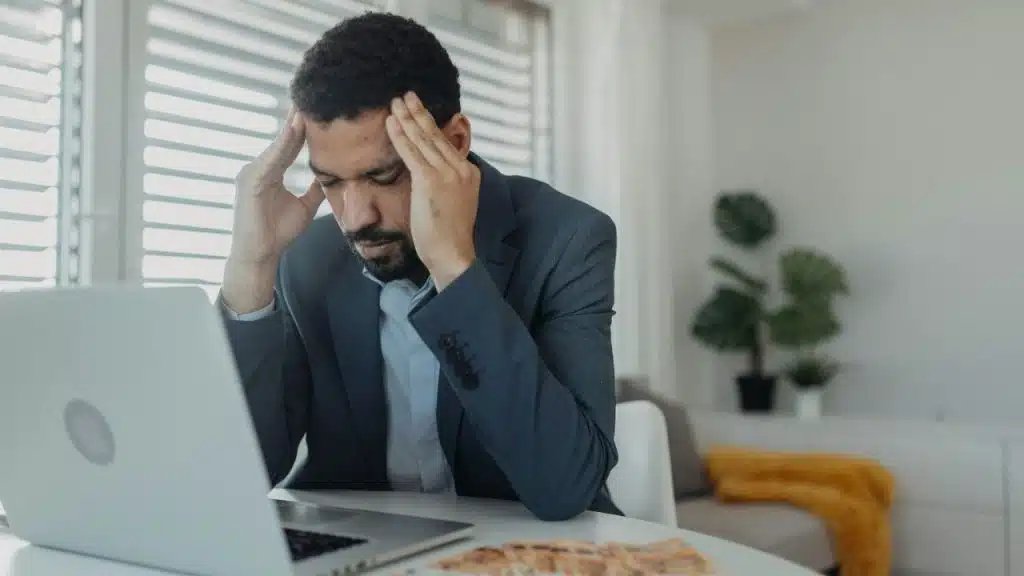 A distressed man with his hands on his head sits at a table with a laptop and money.