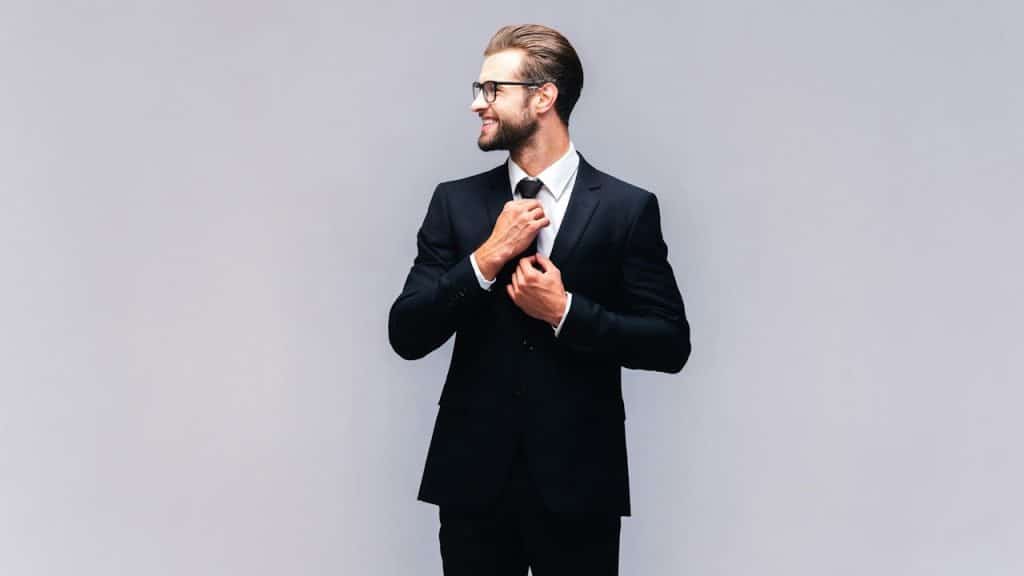 A bearded man wearing glasses and a suit smiles as he adjusts his tie.