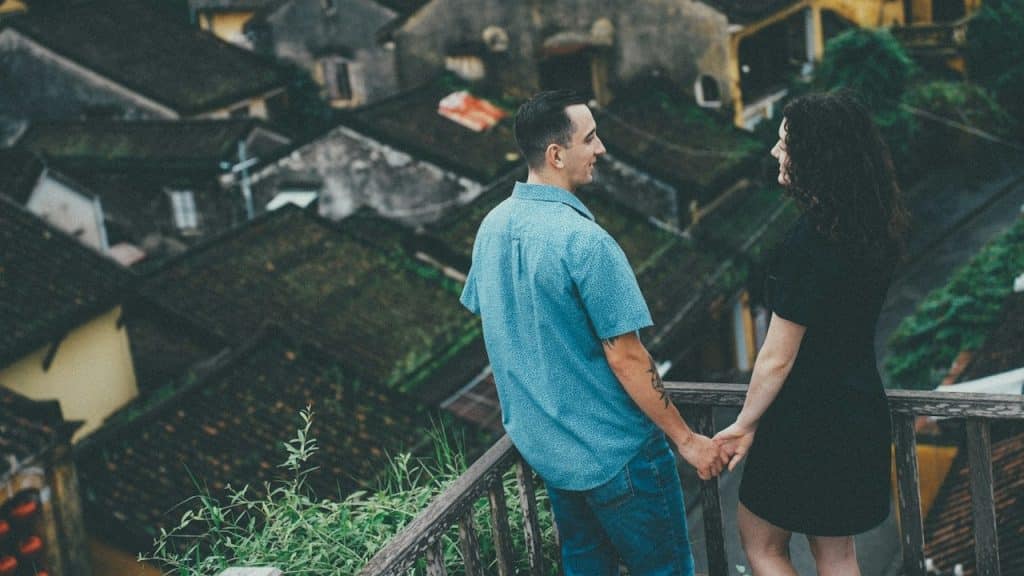 A couple standing on a balcony overlooking a rural area while holding hands.